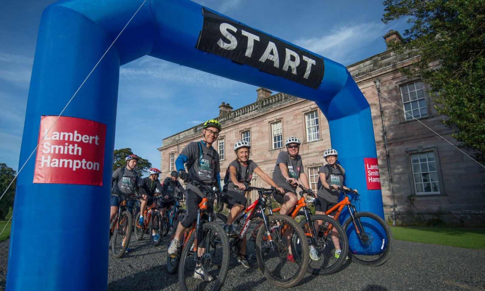 Cyclists grouped together under the start arch before a challenge event.