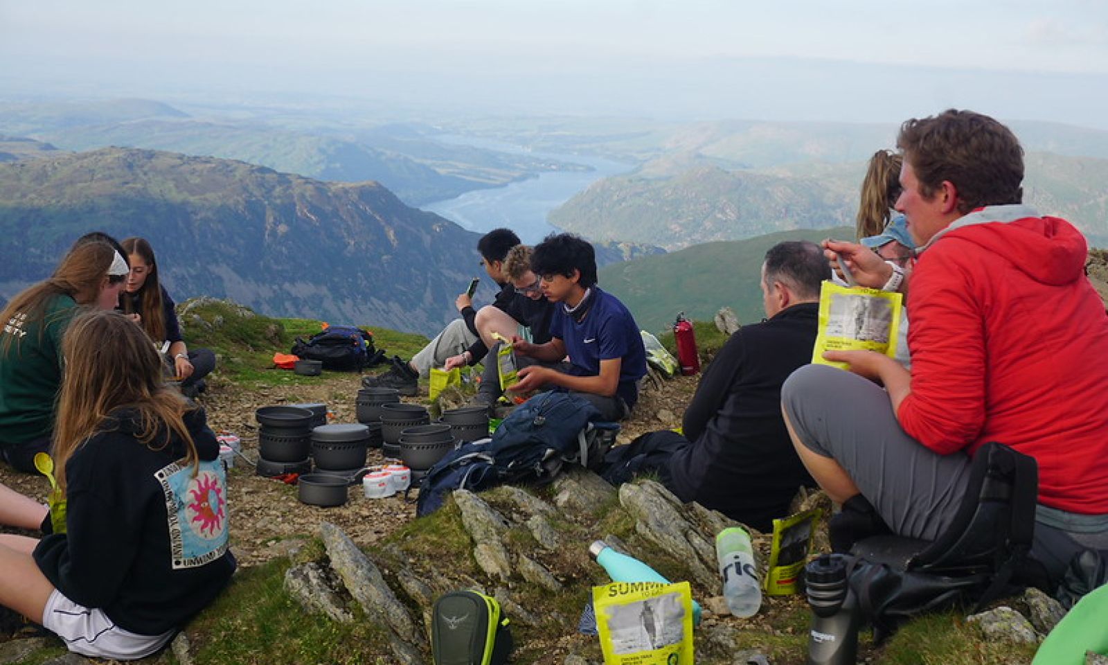 Group of young people sat on the top of a mountain eating their lunch looking out across the mountains